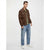 A man modeling a brown utility jacket over a green patterned shirt, paired with blue jeans and white sneakers, against a plain light gray studio background.