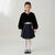 A young girl stands against a light gray studio background wearing a black cropped faux-fur jacket with a single decorative button, a navy pleated skirt, white tights, and black Mary Jane shoes.
