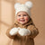 A smiling baby wearing a cream-colored knit beanie with two fluffy pom-poms, matching cream mittens, and a tan winter jacket, set against a warm beige background.