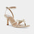 A metallic gold strappy sandal with an ankle strap, a mid-height stiletto heel, and a bow detail on the toe, shown against a light gray background.