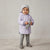 A toddler stands on a light carpet wearing a light lavender quilted hooded jacket, gray leggings, gray boots, and a light gray beanie, while holding a gray stuffed bunny.