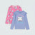 Two children's long-sleeve shirts shown flat on a light gray background: a pink shirt with repeated white cat prints and a light blue shirt with a large white cat graphic and pastel text.