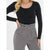 A woman wearing a fitted black long-sleeve scoop-neck top tucked into high-waisted gray paperbag pants with a belt and silver buckle.