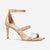 A single nude beige patent leather high-heeled sandal with an ankle strap, buckle closure, and a small gold charm, shown from a side profile on a light gray background.