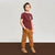 A young boy stands against a neutral studio backdrop wearing a short-sleeve crewneck t-shirt in maroon with a small graphic on the left chest, paired with mustard brown corduroy pants and beige sneakers.