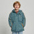 A smiling young boy with light brown hair stands against a light gray studio background wearing a teal hooded puffer jacket with orange piping and zipper accents, a brown shirt underneath, and blue jeans.
