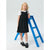 A young girl stands on a small blue step stool in a studio setting, wearing a black pinafore-style dress over a white short-sleeve top, with black loafers and white socks.