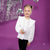 A young girl with blonde hair in two buns wearing a white long-sleeve blouse with ruffle details and black pants, standing against a purple background with silver tinsel and disco ball decorations.