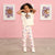 A young girl modeling a cream-colored two-piece outfit featuring a sequined flower graphic on the t-shirt and matching floral print pants, posing in front of a pink wall with framed art.
