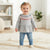 A toddler girl wearing a gray long-sleeve top with a colorful geometric pattern across the chest, blue jeans, and cream shoes, standing on a textured cream blanket in a bright room.