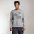 A man wearing a light gray crewneck sweatshirt with a black line-art graphic of a motorcycle engine design and the text 'Motorcycle engine design' on the front, paired with dark gray pants, against a plain light gray studio background.