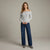 A woman modeling an off-the-shoulder light gray knit sweater and dark blue wide-leg jeans against a neutral studio backdrop.