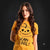 A woman with long wavy brown hair models a yellow graphic t-shirt featuring a black pumpkin illustration and the text 'SCARY COOL!' against a dark studio background.