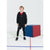 A young boy wearing a black zip-up hoodie, black shorts, and black leggings stands next to a blue and red cube on a white studio floor with colored lines.