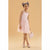 A young girl with her hair in two buns wearing a light pink sleeveless dress and holding a matching light pink handbag, standing against a solid beige background.