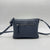 A dark blue leather crossbody bag with a front zippered pocket and a matching shoulder strap, photographed against a neutral gray background.
