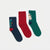 Three Christmas-themed crew socks laid flat on a white background: navy with a tree and lights, solid red, and green with a mummy and baby Jesus design.