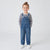 A young child standing against a light gray studio background, wearing blue denim overall with a navy and white striped long-sleeve shirt underneath and white sneakers.