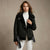 A woman modeling a black shearling-lined aviator jacket with a white turtleneck and white trousers, standing against a neutral textured beige studio backdrop.