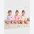 Three babies sitting side by side on a cream knit blanket, wearing long-sleeve baby bodysuits. The baby on the left wears a light purple and white striped bodysuit with a pink flower on the chest.