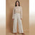 A woman stands against a warm beige studio backdrop wearing a beige ribbed knit long-sleeve top and light beige wide-leg pants with brown sandals.