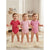 Three babies standing on a fluffy cream rug in a softly lit nursery, each wearing a short-sleeve bodysuit in a different pink shade: a dark raspberry pink, a light bubblegum pink, and a medium pastel pink.