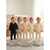 Five toddlers standing together in a softly lit nursery, each wearing a one-piece footed pajama in a different neutral color and pattern: solid black, beige with small dark spots, peach with small dark spots, cream with peach polka dots, and cream with peach stripes.