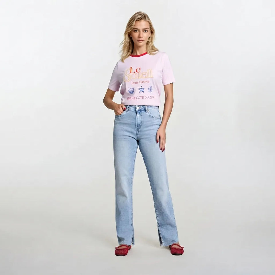 A woman modeling a light pink graphic t-shirt with red trim and light-wash blue jeans, standing against a plain white studio background.