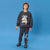 A young boy stands against a solid blue studio background wearing a dark gray sweatshirt and sweatpants set with dinosaur graphics. The sweatshirt features a large T-Rex illustration and the text 'DINOSAUR T-REX', while the pants have a matching graphic on the lower leg.