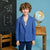 A young boy with curly brown hair stands in a classroom-like setting wearing a bright blue blazer and navy pants. A chalkboard with white chalk writing is on the left, and a globe on a wooden table is on the right.