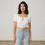 A woman modeling a white short-sleeve crop top with lace trim and a front tie closure, paired with light-wash blue jeans, against a plain off-white studio background.