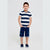 A young boy stands against a light gray studio background wearing a navy and white horizontally striped short-sleeve t-shirt, dark blue denim shorts, and navy slip-on shoes.