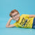 A young boy with blond hair and round black-rimmed glasses lies on his side against a solid light blue background, wearing a bright yellow SpongeBob-themed t-shirt and blue jeans.