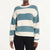 A woman wearing a ribbed knit sweater with wide horizontal stripes in teal blue and off-white, paired with black pants, against a plain white studio background.