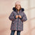 A young girl wearing a dark floral print winter parka with a dark faux-fur hood trim, a pink knit beanie, and blue jeans, standing against a soft pink background.