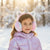 A young girl wearing pink and white fuzzy unicorn earmuffs with a silver horn, paired with a shiny lavender puffer jacket, standing in a snowy outdoor setting with warm sunlight and bokeh.