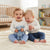Two smiling toddlers sitting on a cream knit blanket in a bright nursery, wearing matching white long-sleeve tops and light blue leggings. One child holds a soft blue stuffed bunny.