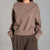 A person wearing a taupe-brown crewneck sweatshirt and dark brown pleated trousers, standing against a neutral gray studio background.