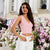 A woman modeling a light pink ribbed knit wrap-style crop top and white pants, standing outdoors in front of a background of pink and white flowers.