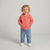 A toddler with curly blonde hair stands against a light gray studio background wearing a coral pink zip-up hooded jacket, blue jeans, and white sneakers. The jacket has a small embroidered logo on the left chest.