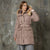 A woman wearing a taupe-brown puffer coat with a faux-fur trimmed hood and a matching belt, standing against a textured gray concrete wall.