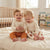 Two smiling toddlers sitting on a cream ribbed blanket in a bright nursery. The child on the left wears a cream long-sleeve top and rust-orange pants, holding a small teddy bear.