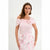 A woman modeling a light pink off-the-shoulder top with a large fabric rose detail on the chest, paired with a pink and white floral skirt, against a plain light gray background.