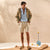 A man stands in a studio wearing a light brown jacket over a light blue button-down shirt and beige cargo shorts, paired with tan sneakers. The background is a warm beige textured wall with soft shadows, and a small blue decorative stand with yellow spheres sits to the side.