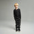 A young boy with blonde hair stands against a neutral gray studio background wearing a black long-sleeve henley shirt with three gold buttons, dark gray textured jogger pants, and black sneakers.