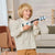 A young blonde boy wearing a beige and white striped long-sleeve shirt and tan pants is holding and playing a light blue wooden ukulele in a brightly lit living room.
