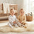 Two toddlers sitting on a fluffy cream rug in a bright nursery. The child on the left wears a white footed one-piece with a small animal print and holds a beige stuffed bunny.