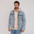 A man wearing a light-wash blue denim jacket over a beige hoodie and blue jeans, standing against a plain light gray studio background.