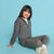 A young girl with dark hair sits smiling on a solid light blue background, wearing a dark gray zip-up hoodie, matching gray joggers, white socks, and chunky off-white sneakers.