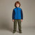 A young boy with curly red hair stands against a neutral studio background wearing a blue and navy zip-up fleece jacket, olive green cargo pants, and green sneakers.